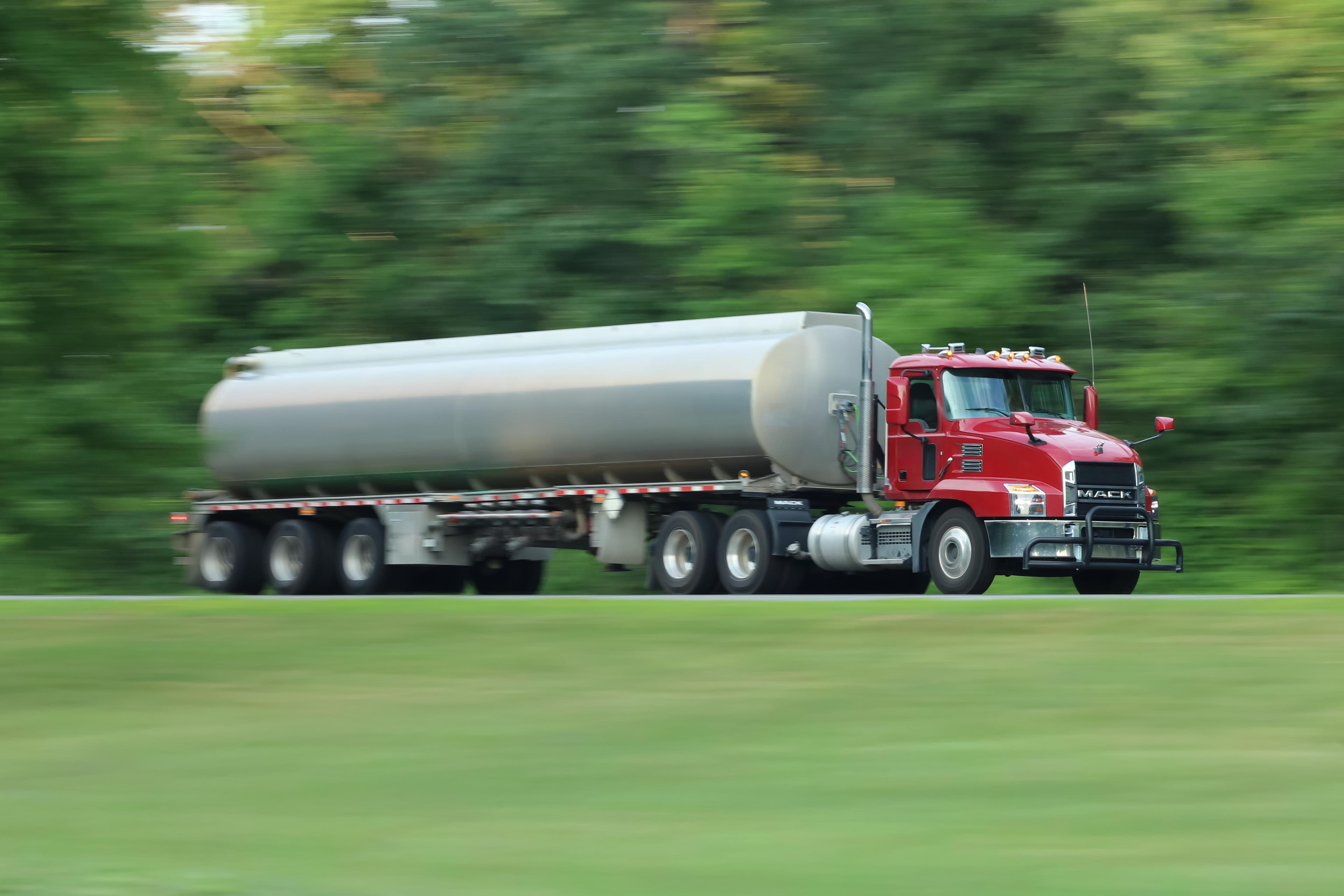 Red Mack truck hauling a silver tanker trailer traveling on highway with motion blur background showing trees and green landscape