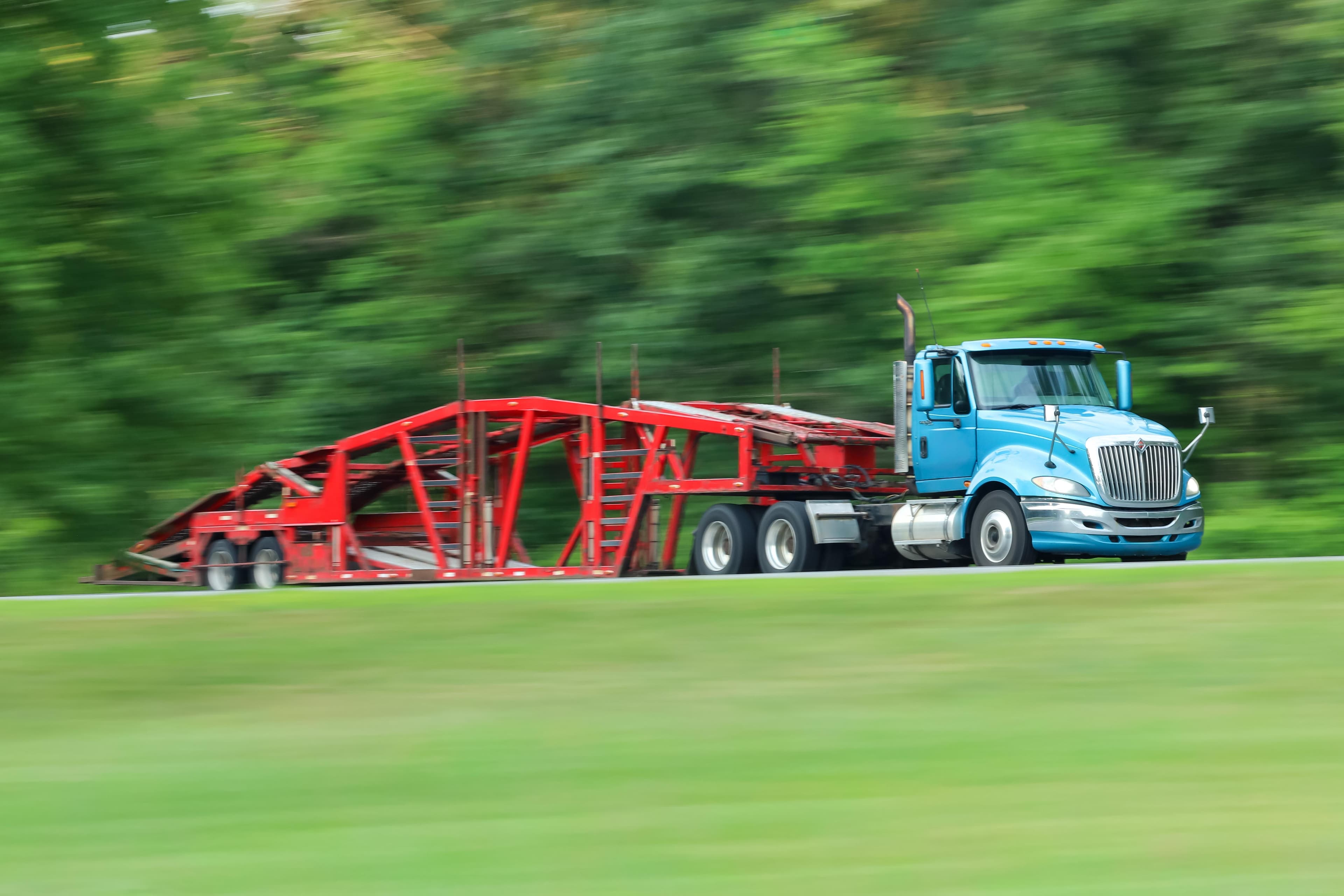 Blue semi-truck pulling a red car carrier trailer on a highway with motion blur effect showing speed and lush green trees in background