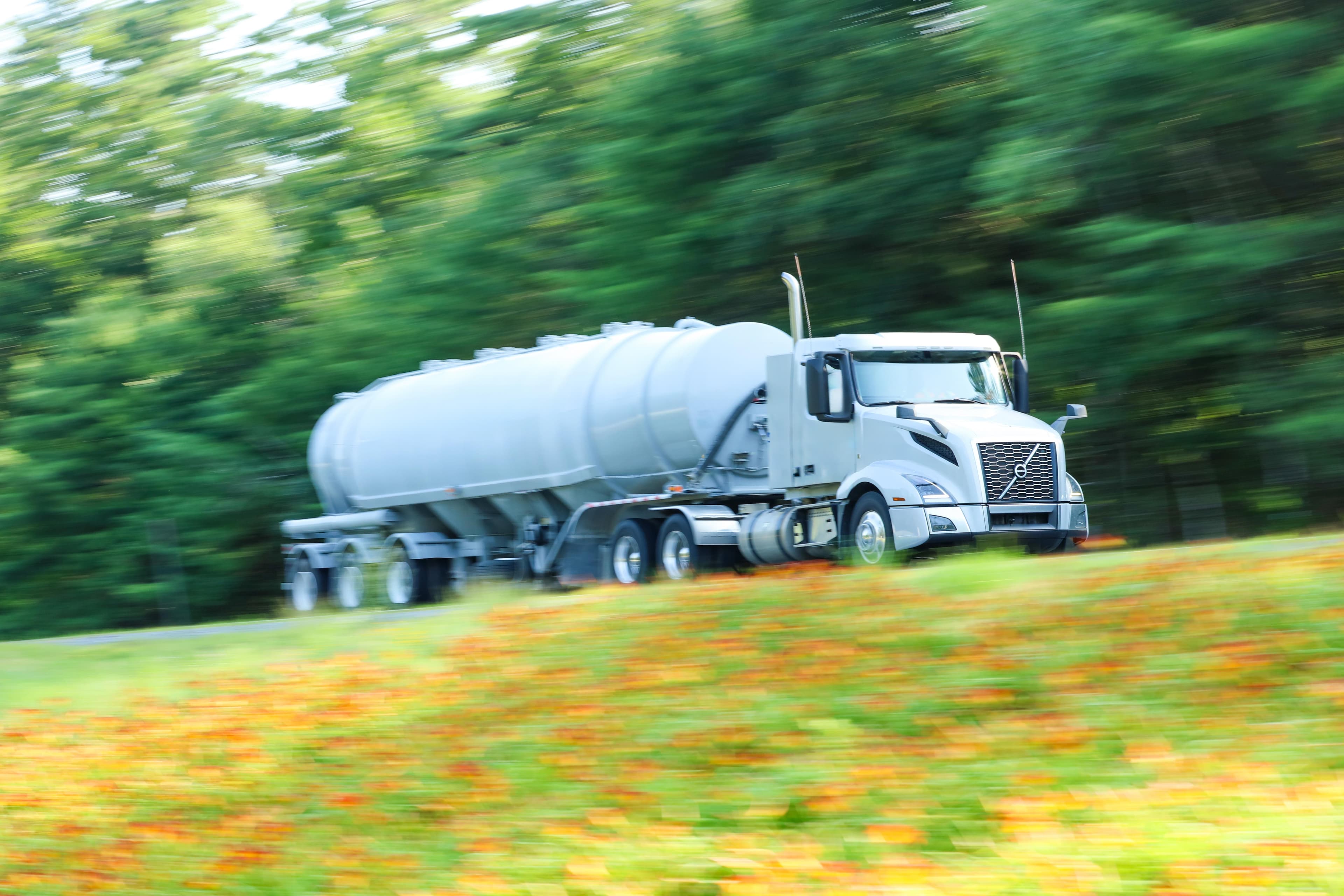 White Volvo truck pulling white tanker trailer on rural road with motion blur effect from camera panning, surrounded by lush green foliage