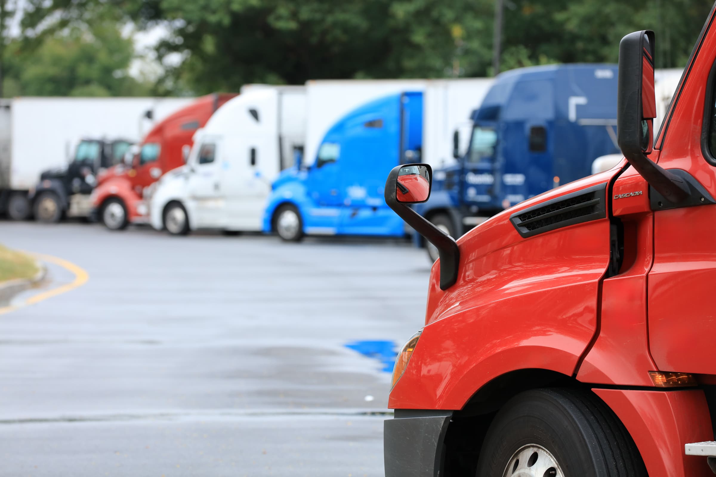 Close-up view of a red semi-truck cab in the foreground with multiple commercial trucks parked in the background at what appears to be a truck stop or rest area