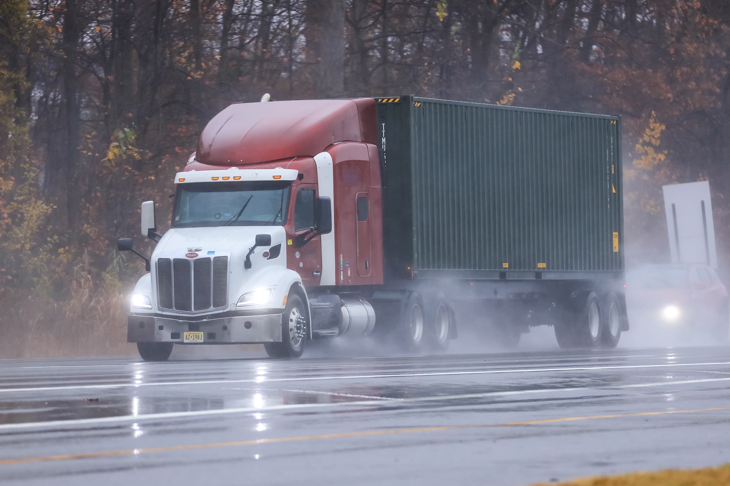 Peterbilt semi-truck with white cab and red sleeper pulling a green shipping container on wet highway during rainy autumn conditions with trees in background