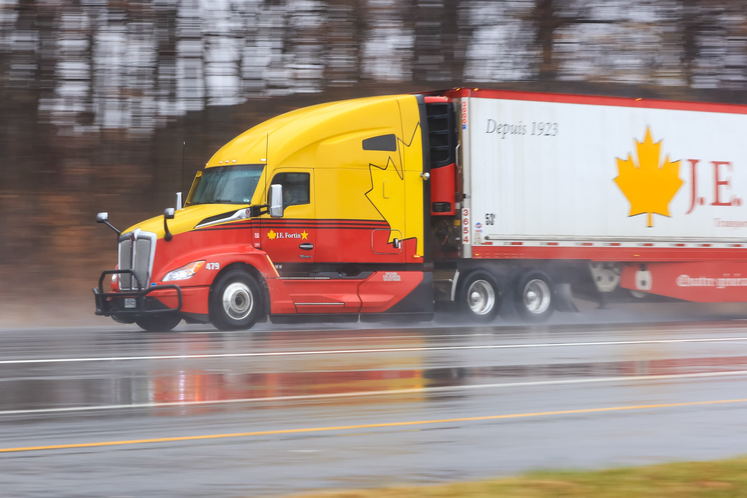 Red and yellow Peterbilt truck pulling white J.E. branded trailer on wet highway with motion blur